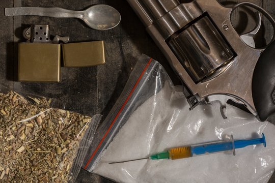 Hard drugs and gun revolver on dark table. A dark theme, drug use. White powder, marijuana, heroin, hard drugs, lighter and a spoon with pills.