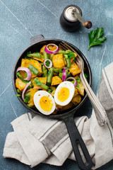 Warm potato salad with green beans, pepper, parsley, eggs and red onion in frying pan on gray concrete background. Selective focus. Top view. Copy space.