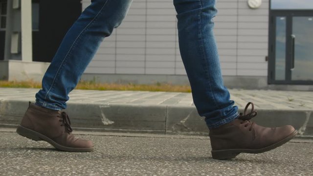 The one guy slowly confidence walks along the town footway in blue jeans and stylish leather shoe. Point of view on legs closeup. Spring cold weather, pedestrian goes past the building slow motion