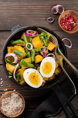 Warm potato salad with green beans, pepper, parsley, eggs and red onion in frying pan on old wooden background. Selective focus. Top view. Copy space.