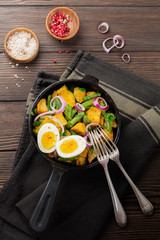 Warm potato salad with green beans, pepper, parsley, eggs and red onion in frying pan on old wooden background. Selective focus. Top view. Copy space.
