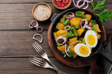 Warm potato salad with green beans, pepper, parsley, eggs and red onion in frying pan on old wooden background. Selective focus. Top view. Copy space.