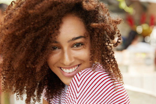 Glad cheerful African American female model with frizzy dark bushy hair, being in high spirit after hearing good news or recieving compliment from boyfriend, poses at camera with gentle smile.
