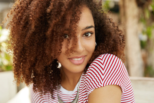 Close up shot of lovely dark skinned mixed race female with wavy dark hair, smiles pleasantly, wears striped t shirt, being satisfied with everything. Adorable African woman expresses happiness