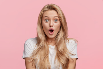 Blue eyed blonde young woman in casual white t shirt, looks with opened mouth, being shocked to recieve award after participating in competition, isolated over pink background. Shock concept - obrazy, fototapety, plakaty