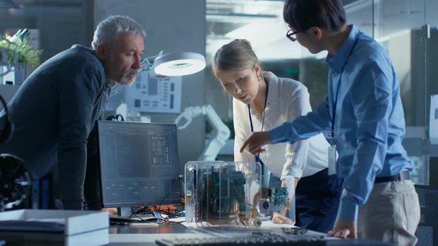 Team of Computer Engineers Lean on the Desk and Choose Printed Circuit Boards to Work with, Computer Shows Programming in Progress. In The Background Technologically Advanced Research Center.