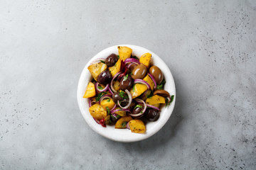 Warm potato salad with olives, pepper, parsley and red onion on old white ceramic plate on gray concrete background. Selective focus. Top view. Copy space.
