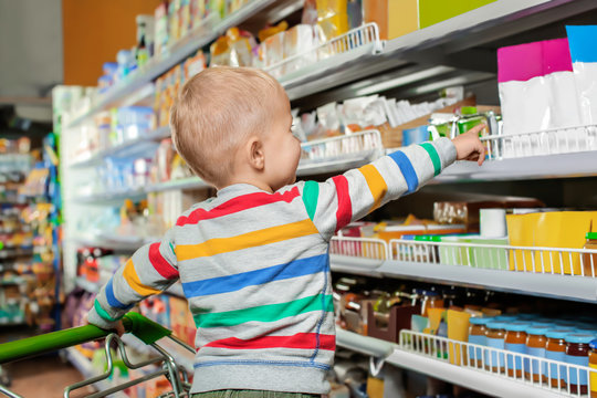 Cute little boy in shopping cart choosing baby food at supermarket