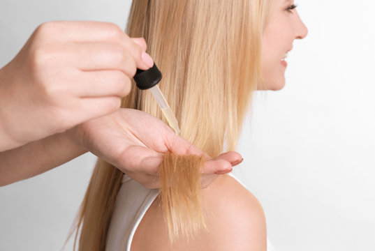 Stylist applying oil onto woman's hair on light background