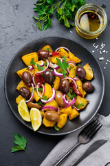 Warm potato salad with olives, pepper, parsley and red onion on black ceramic plate on dark concrete background. Selective focus. Top view. Copy space.