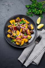 Warm potato salad with olives, pepper, parsley and red onion on black ceramic plate on dark concrete background. Selective focus. Top view. Copy space.