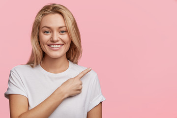 Pretty young blonde woman with positive expression, dressed casually, indicates with fore finger at...