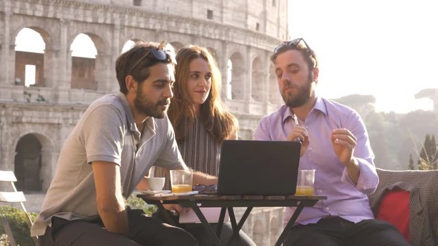 Three young people having job videocall with laptop working outside together pitching a project sitting at bar restaurant table in front of colosseum in rome at sunset
