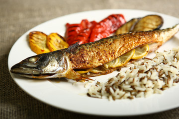 Healthy and proper food, grilled fish and vegetables with lemon and black and white rice on a white plate. Against the background of sacking and a wooden table. Low-calorie food