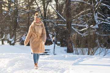 Young caucasian woman walking on snow path in park at sunny frosty day, enjoying winter moments and nature, wearing winter clothes - beige coat, scarf, hat with pompon/ Winter time, walking - concept 