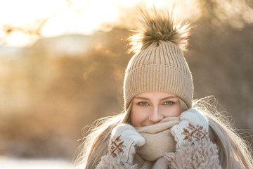 Portrait of young European woman with long hair in beige hat with fur pompon, scarf, coat, white gloves at sunny winter day/ Female wrap in and holding scarf, smiling, looking at camera, copy space