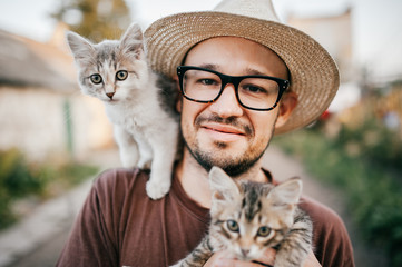 Happpy young bearded farmer holding two little kitten in hands outdoor in village with abstract background. Smiling man in glasses and straw hat playing with funny cute pets. Have fun in countryside.