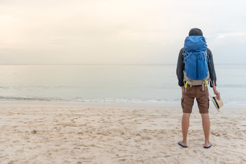 Young Asian backpacker man standing on the beach and holding hat, summer holiday vacation and travel tropical island concepts