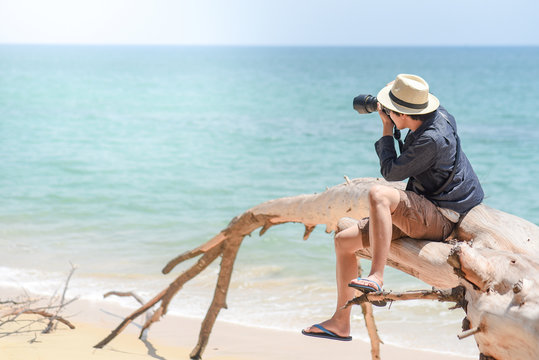 young man photographer with jean shirt and hat sitting on white dead tree near sandy beach of tropical island and taking photo of turquoise sea, summer holiday and vacation travel concepts
