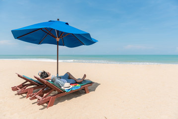 Young Asian man traveler lying on beach bench with blue umbrella overhead. surrounded by beautiful tropical beach island and blue sky, vacation time and summer holiday for digital nomad lifestyle