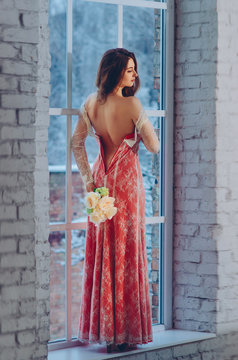 Girl in long red dress stands before white window holding white bouquet before her naked back