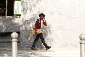Obraz na płótnie Canvas Full body african guy with skateboard walking outside using mobile phone