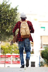 behind of young man walking outdoors with bag and skateboard
