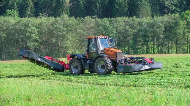 A red tractor is pulling two machines and it is cutting grass. The day is beautiful and sunny.