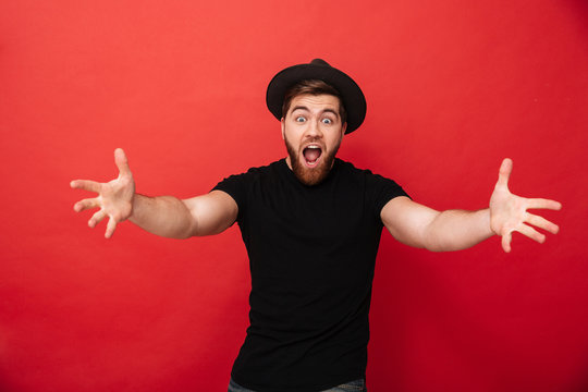 Photo of funny happy man wearing black t-shirt and hat emotionally throwing hands up on camera, isolated over red background
