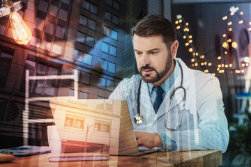 Smart doctor. Calm attentive serious doctor looking thoughtful while sitting at the table and reading the news on the screen of his laptop
