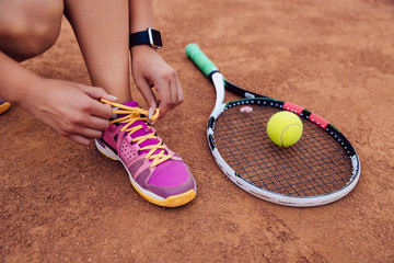 Athlete woman getting ready for playing a game of tennis, tying shoelaces. Close-up view of racket...