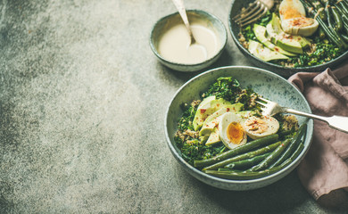 Healthy vegetarian breakfast bowls. Quinoa, kale, green beans, avocado, egg and creamy tahini dressing bowls over grey concrete background, selective focus, copy space. Energy boosting food concept