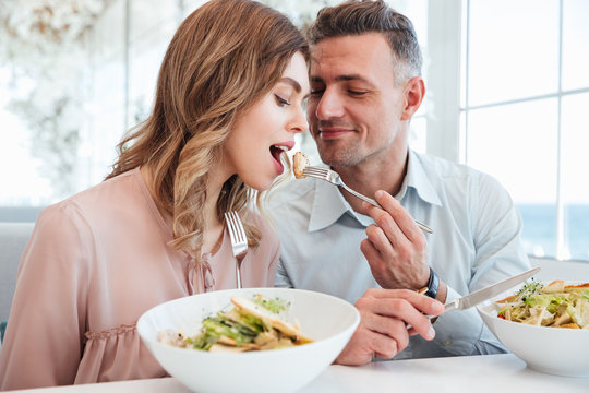 Handsome mature man feeding beautiful woman with salad, while having romantic meal in restaurant on sunny day