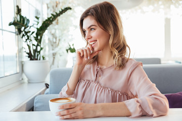 Photo of beautiful smiling woman sitting alone in restaurant in morning, while drinking cappuccino from cup