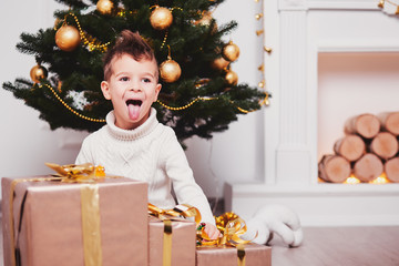 Cute little boy open his mouth and showing tongue near a lot of gifts with golden ribbons and bows....