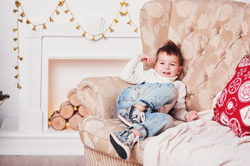 Fototapeta na wymiar A cute little boy laughs and lies on the couch. A cheerful guy in a knitted sweater and a stylish hairdo rests on the couch with his legs tucked. Near the red pillows with a Christmas pattern.
