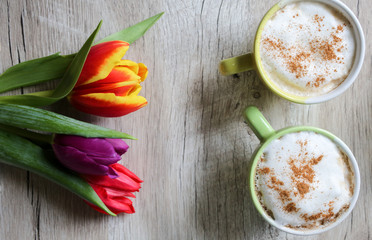 Tulips on wooden background with two cups of coffee. Invitation postcard for mother's day or international women's day. Punchy pastels. Cappuccino with foam cinnamon and flowers.