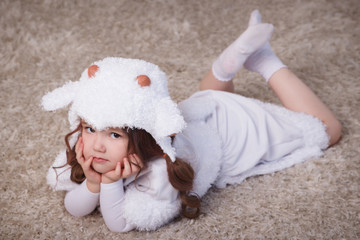 A little girl in her room, lying on the carpet, on the floor, in a lamb suit. Child in white dress.