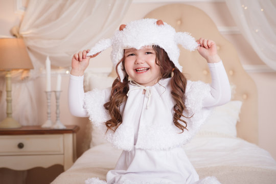 A little girl in her room sits on a bed, in a lamb suit. Child in white dress. Beige interior.