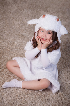 A little girl in her room sits on a carpet, on the floor, in a lamb suit. Child in white dress. The face is frightened, not understanding.