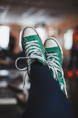 Authentic picture of streched out and crossed female legs wearing with old green sneakers with untied laces, softly blurred old gym on background.