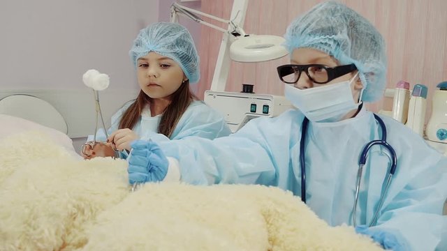 cute and funny children play doctor, boy and girl perform surgery on a bear in the playroom, the nurse gives a clamp with a bandage, and the surgeon operates