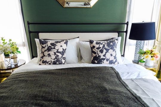 An interior picture of a bed with many pillow with a black blacket and flower bouquet in a pot and luxury lamp.