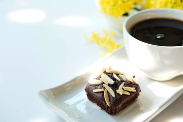 Chocolate brownie cake in white plate on old white table , there is hot coffee in white cup and beautiful yellow flower in the background