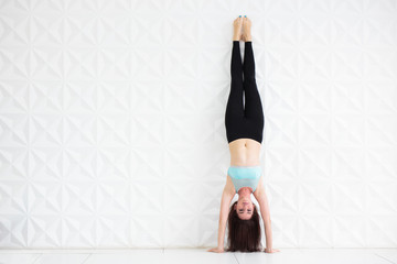Young brunette woman doing a handstand over a white wall