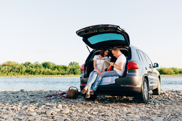 Cheerful couple sitting in car open trunk and watching map, roadtrip concept