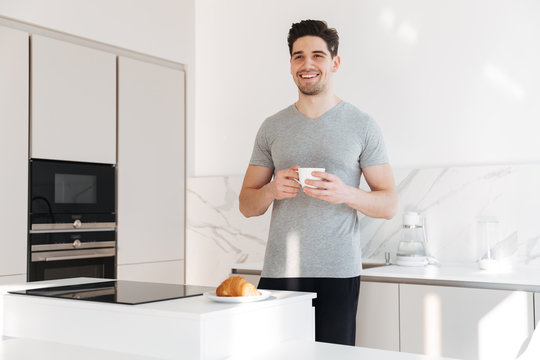 Portrait of adult man in casual clothes smiling and holding cup of coffee, while having breakfast in apartment