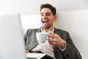 Photo of happy man in businesslike clothes bursting into laughter, while resting in bed with laptop and drinking coffee