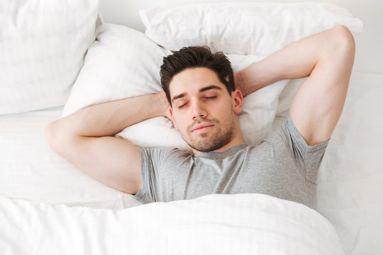 View from above of brunette muscular man in casual t-shirt, sleeping alone at home in bed with white clean linen putting hands under head