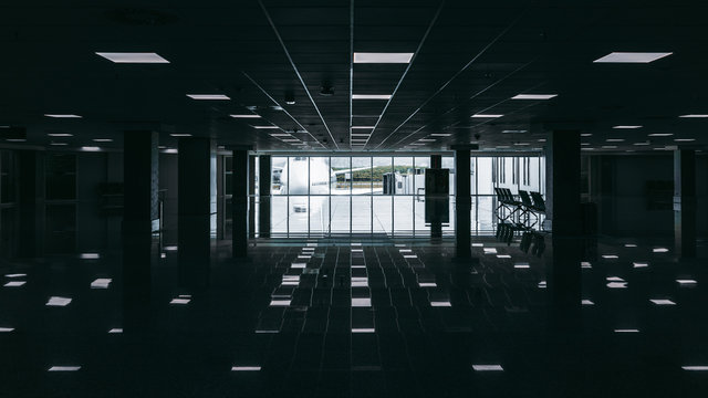 Dark modern airport waiting hall with multiple overhead illumination lamps with reflections stretching into vanishing point, huge window at the end of hall with white double-decker airplane outside